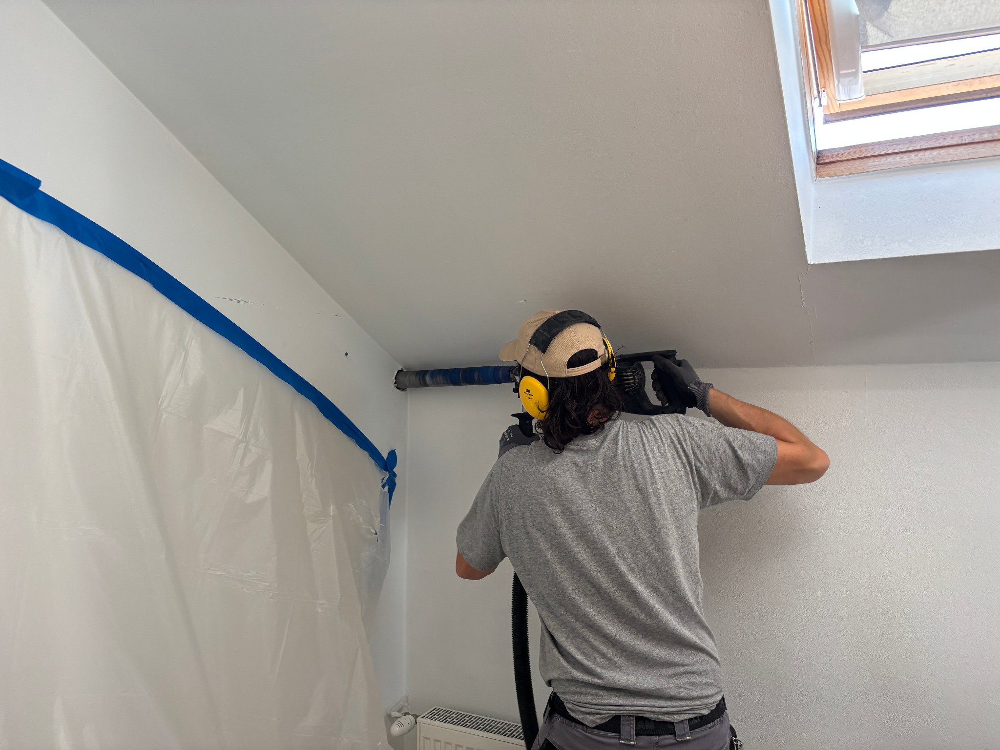 Instalator wierci otwór w ścianie pod klimatyzację, zabezpieczonej folią i taśmą. Widoczne okno dachowe, grzejnik i osłona słuchu.