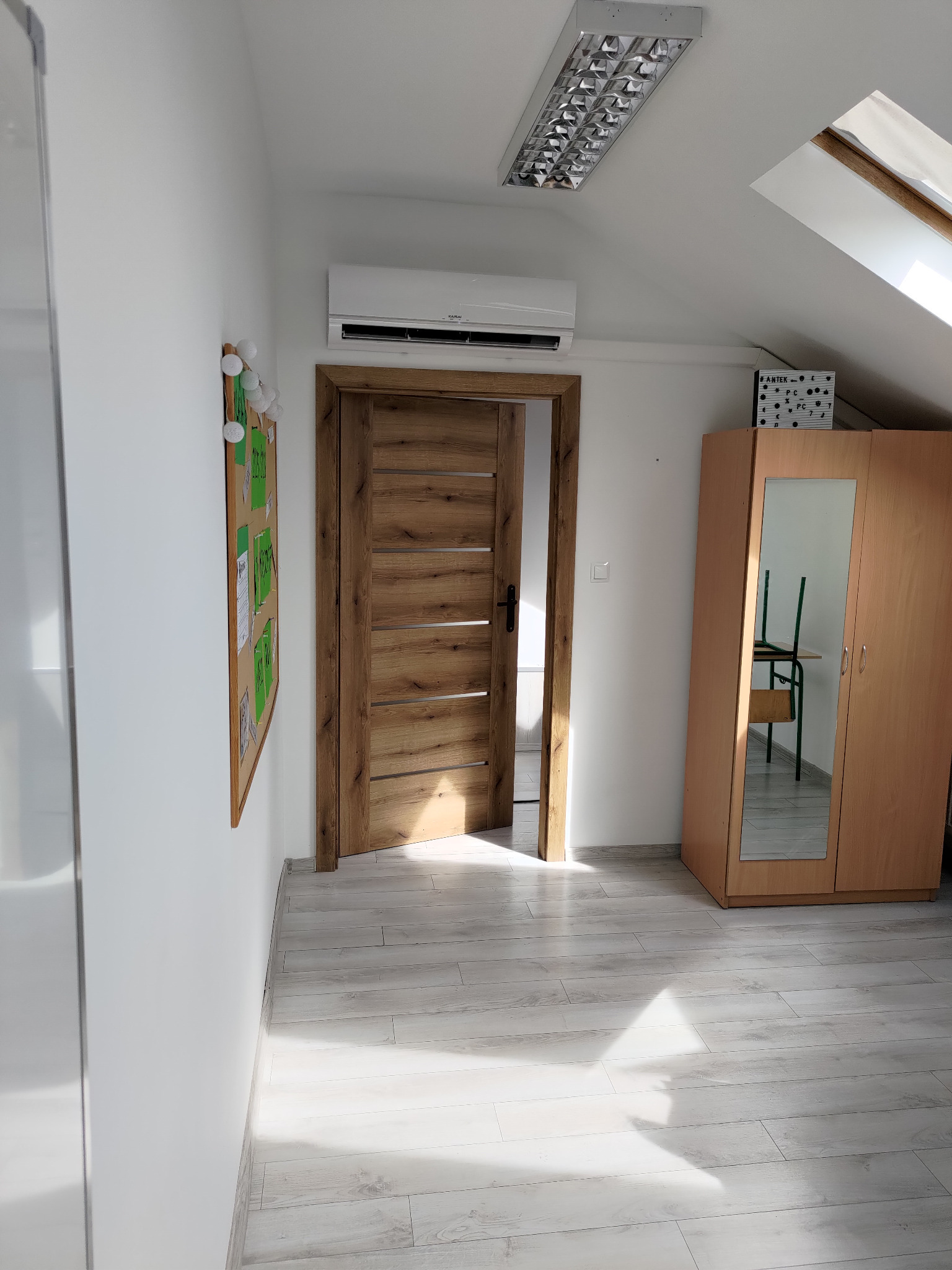 Biały klimatyzator zamontowany w pokoju na poddaszu z oknem dachowym, obok drewnianych drzwi i szafy z lustrem. Widoczna lampa sufitowa i podłoga z paneli.