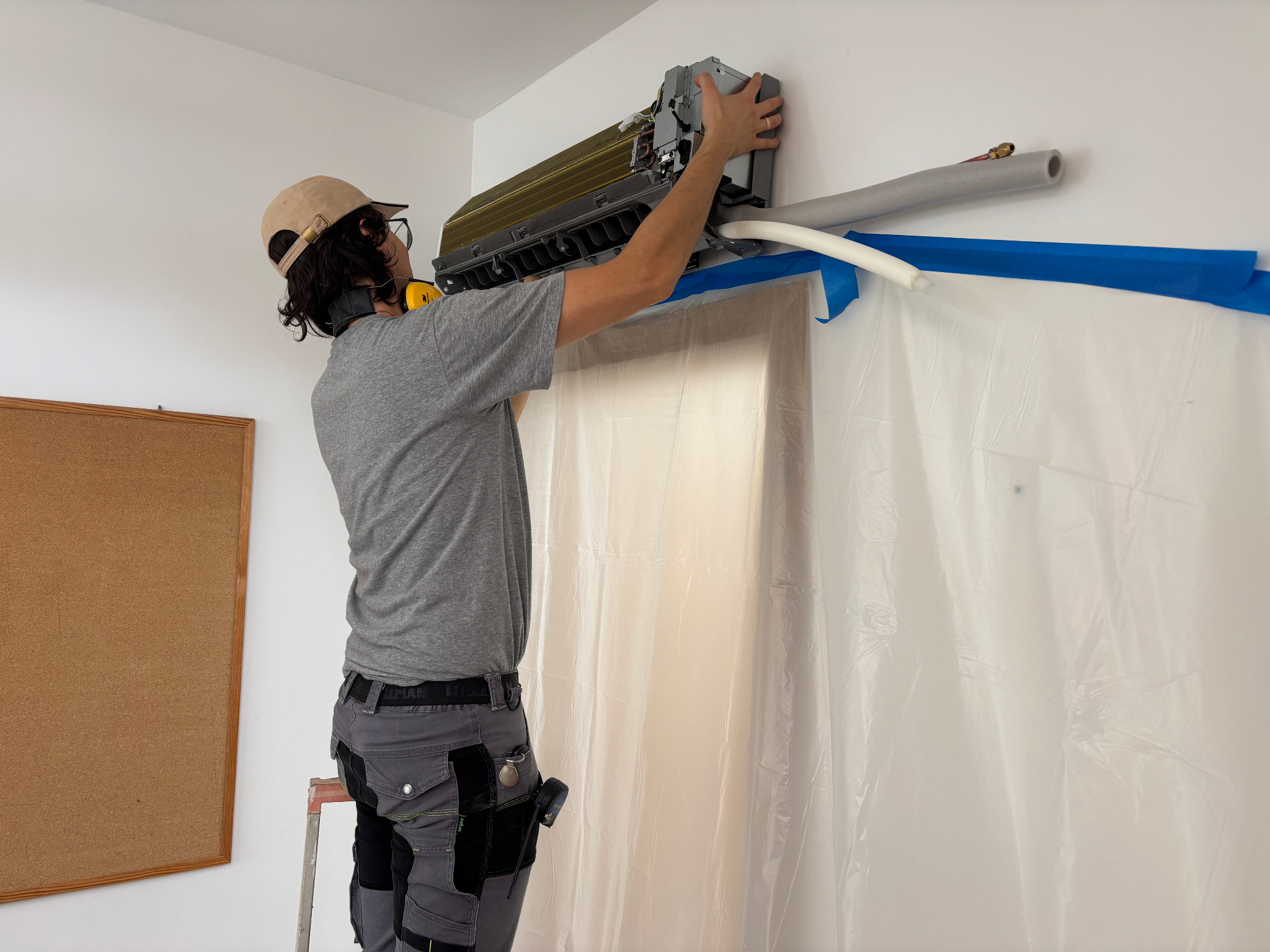 Instalator w czapce montuje klimatyzator na ścianie, widoczne rury i folia ochronna. Praca na wysokości, drabina. Szare spodnie robocze i słuchawki ochronne.