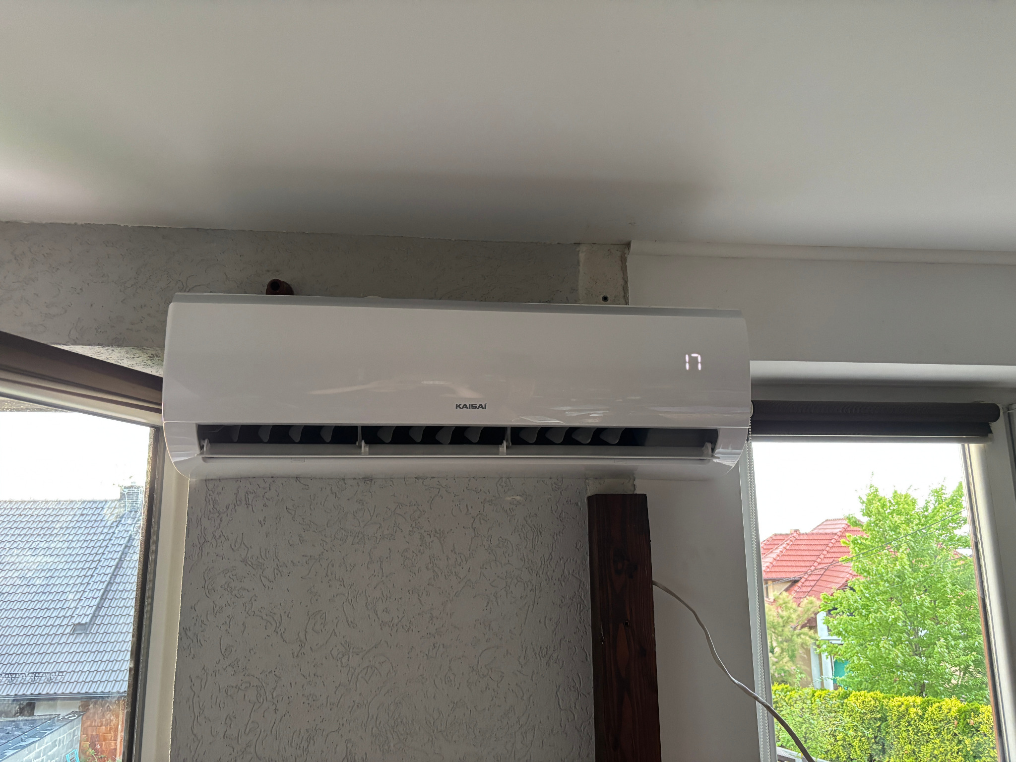 Biały klimatyzator Kaisai zamontowany na ścianie z fakturą, widoczny wyświetlacz z temperaturą 17 stopni, okno z widokiem na zieleń w tle.