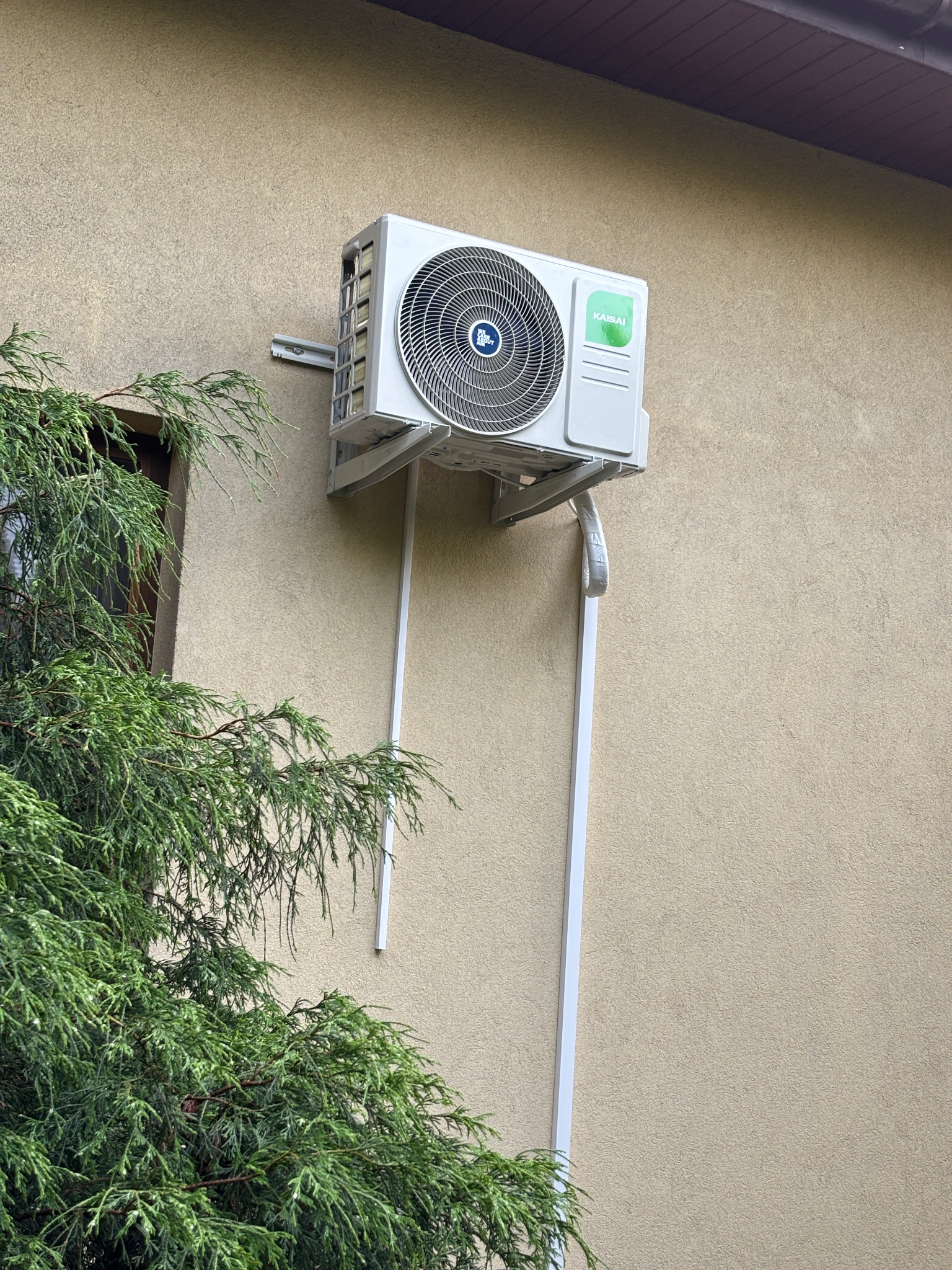 Zamontowany klimatyzator Kaisai na elewacji budynku z widoczną instalacją odprowadzającą skropliny i fragmentem zielonego drzewa iglastego po lewej stronie.