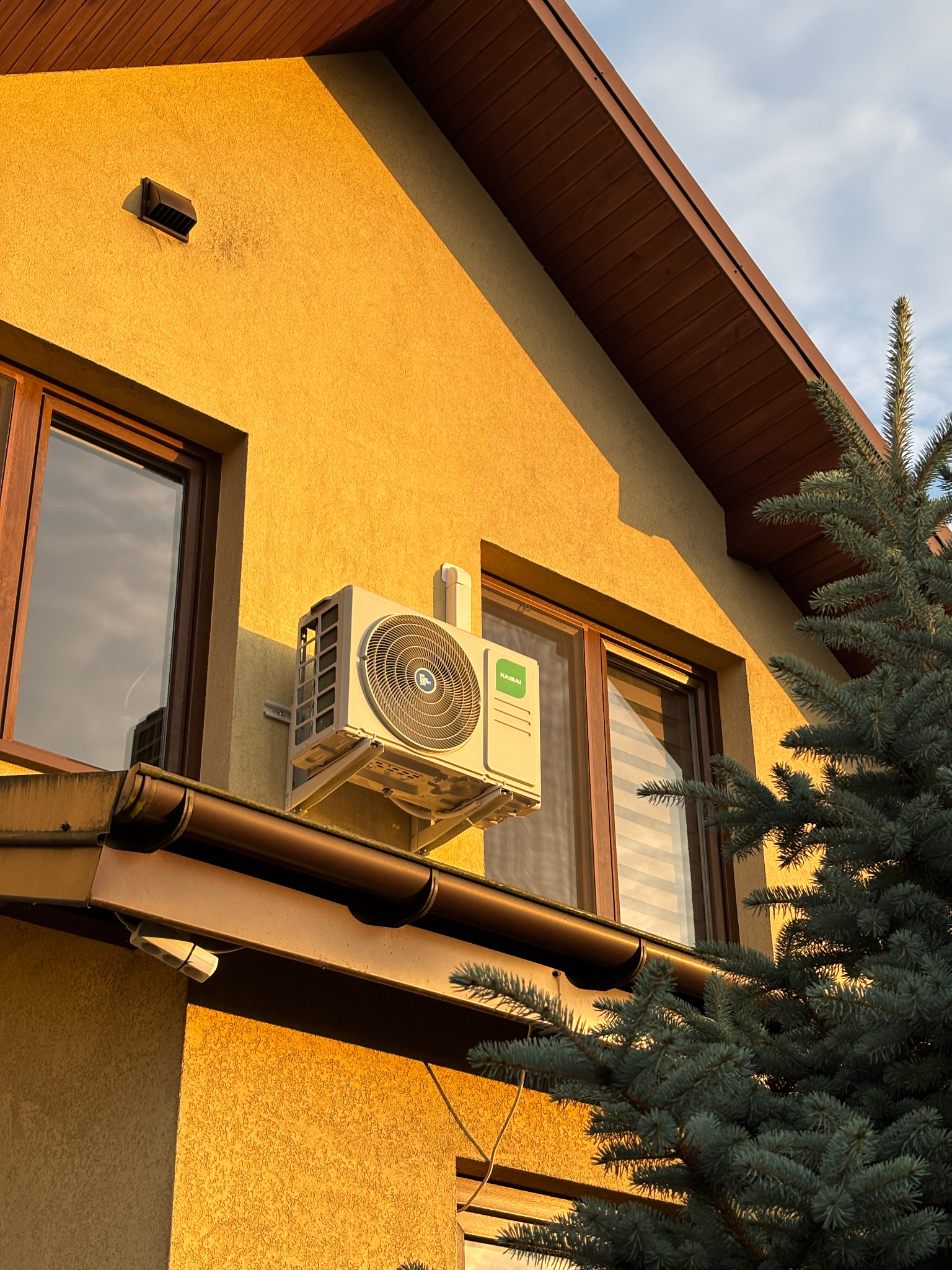 Kompaktowa klimatyzacja marki Kaibai zamontowana na elewacji domu w ciepłym, popołudniowym świetle, obok okna z brązową ramą i fragmentem iglastego drzewa.