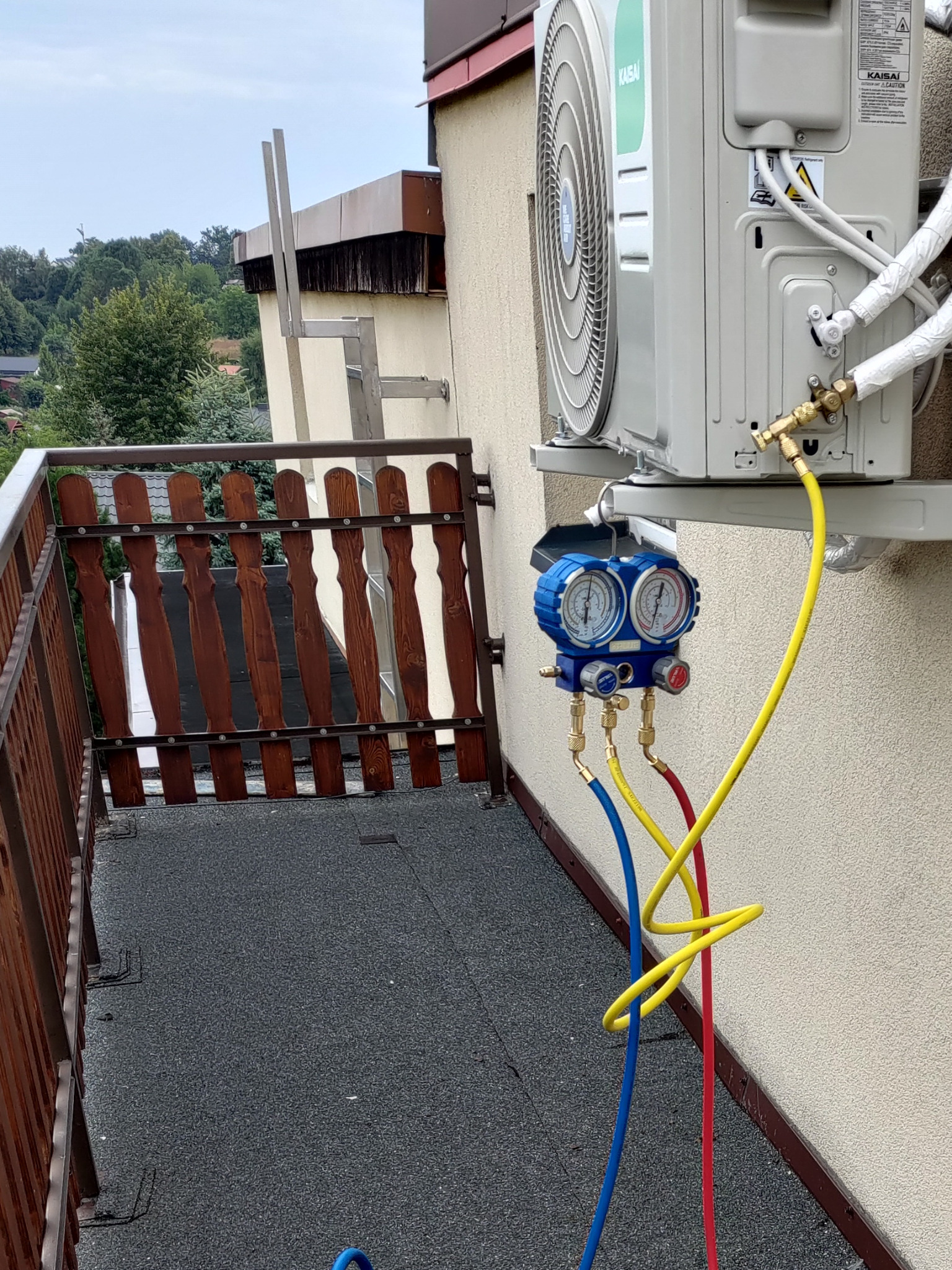Serwis klimatyzacji Kaisai na balkonie: podłączone manometry, widoczne przewody w kolorach żółtym, niebieskim i czerwonym. Zewnętrzna jednostka klimatyzatora na ścianie.