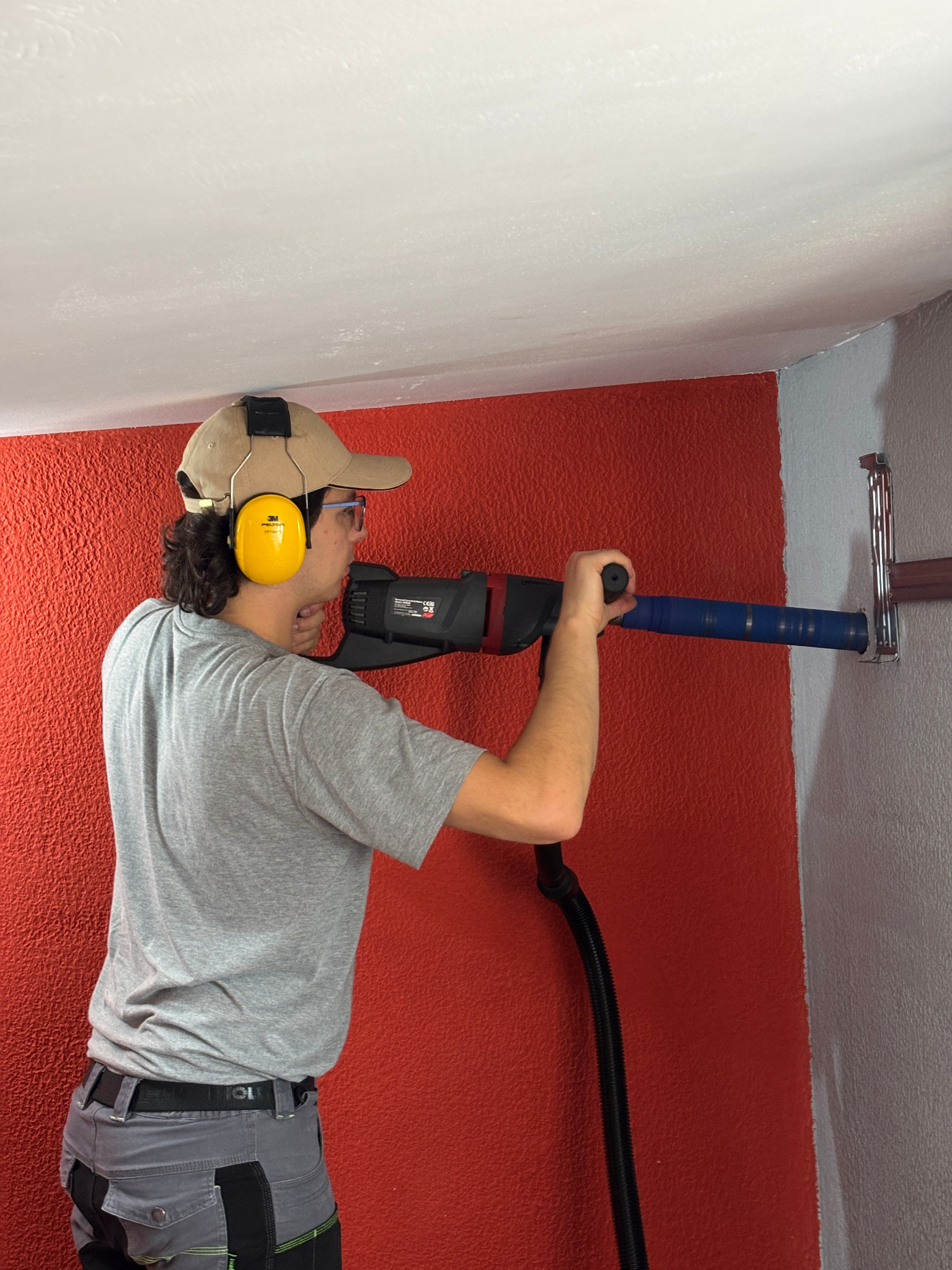 Mężczyzna w słuchawkach wierci otwór w ścianie za pomocą wiertnicy z odsysaniem pyłu. Widoczny uchwyt montażowy. Czerwona ściana, biały sufit.