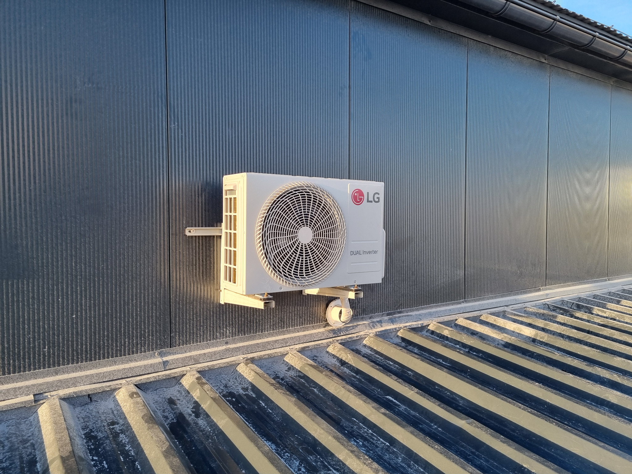 Zamontowana jednostka zewnętrzna klimatyzacji LG Dual Inverter na ciemnej, metalowej ścianie budynku z widocznym fragmentem dachu. Ujęcie z dołu.