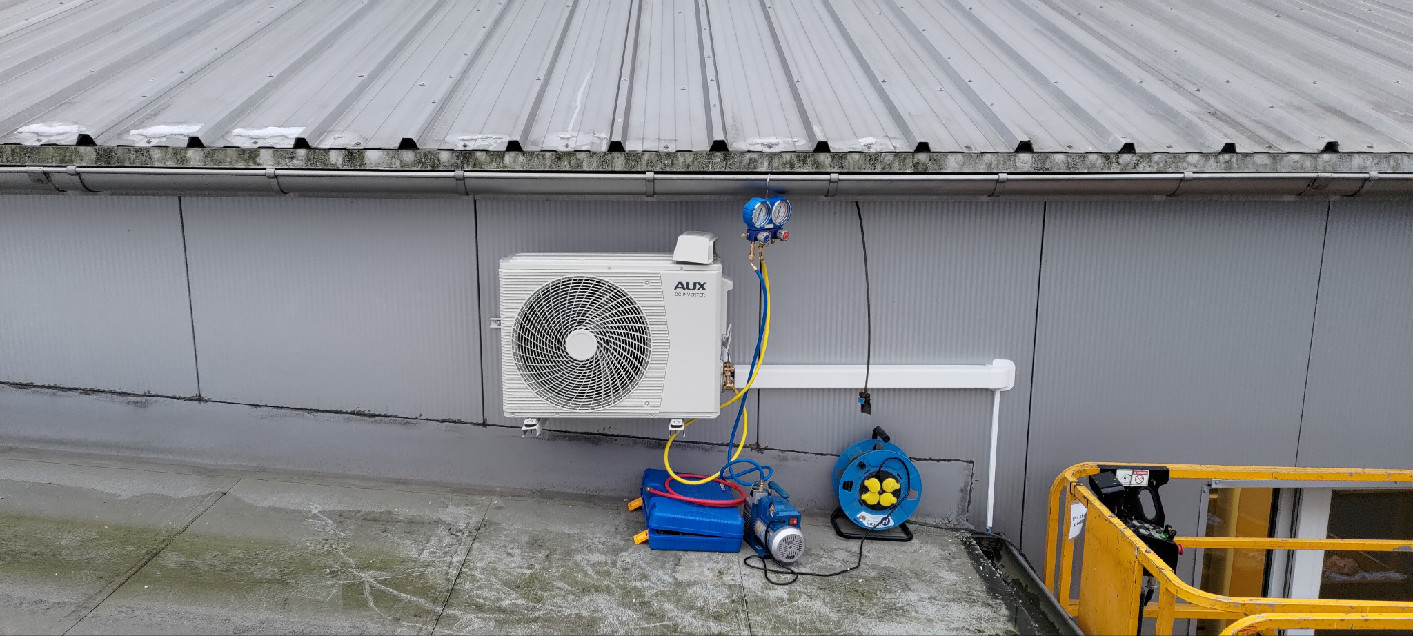 Biały klimatyzator AUX DC Inverter zamontowany na szarej ścianie budynku z blachy trapezowej. Podłączone narzędzia serwisowe, widoczny fragment podnośnika.