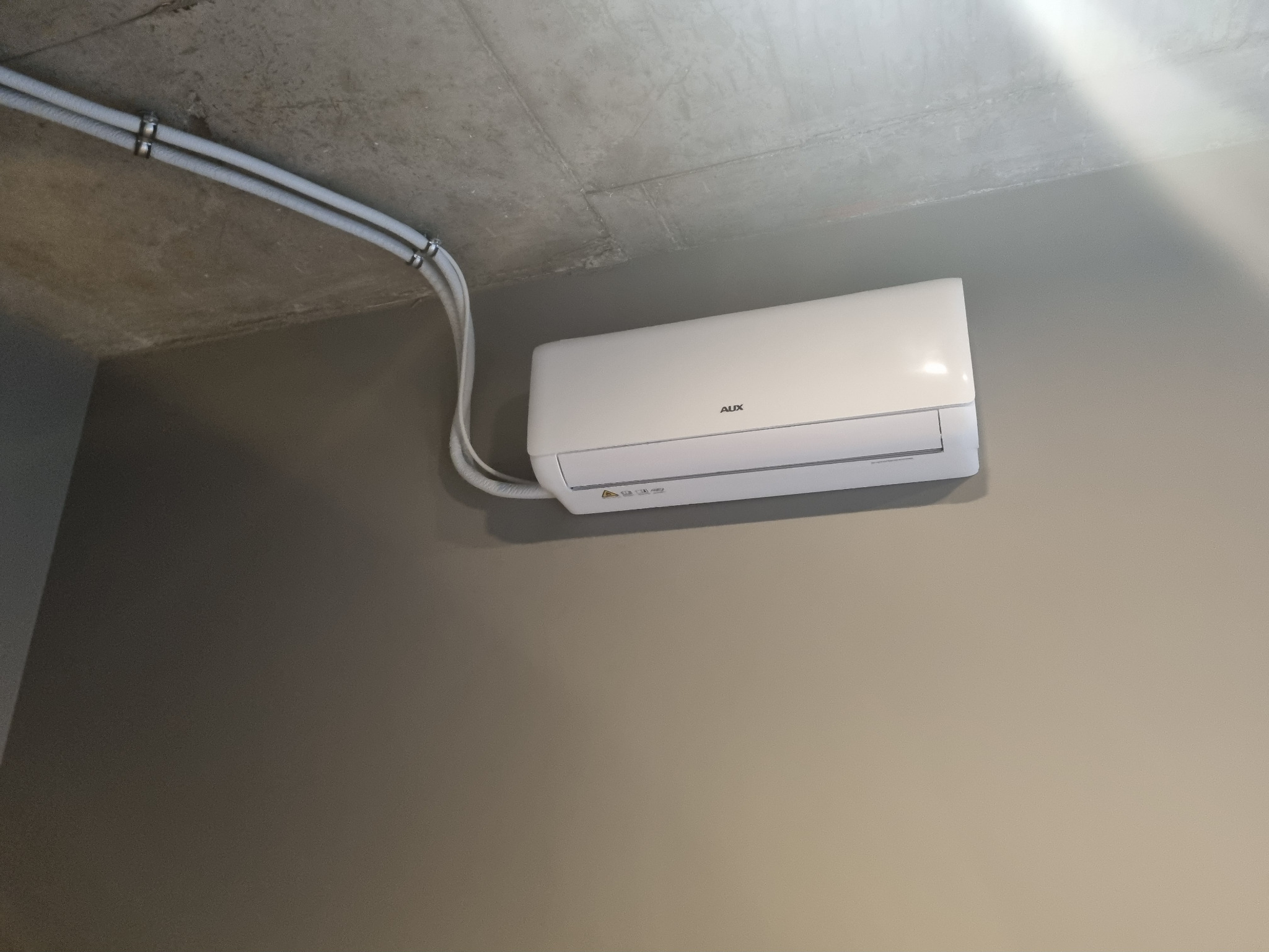 Biały klimatyzator AUX zamontowany na szarej ścianie, widoczne rury instalacyjne przymocowane do sufitu. Minimalistyczny design.