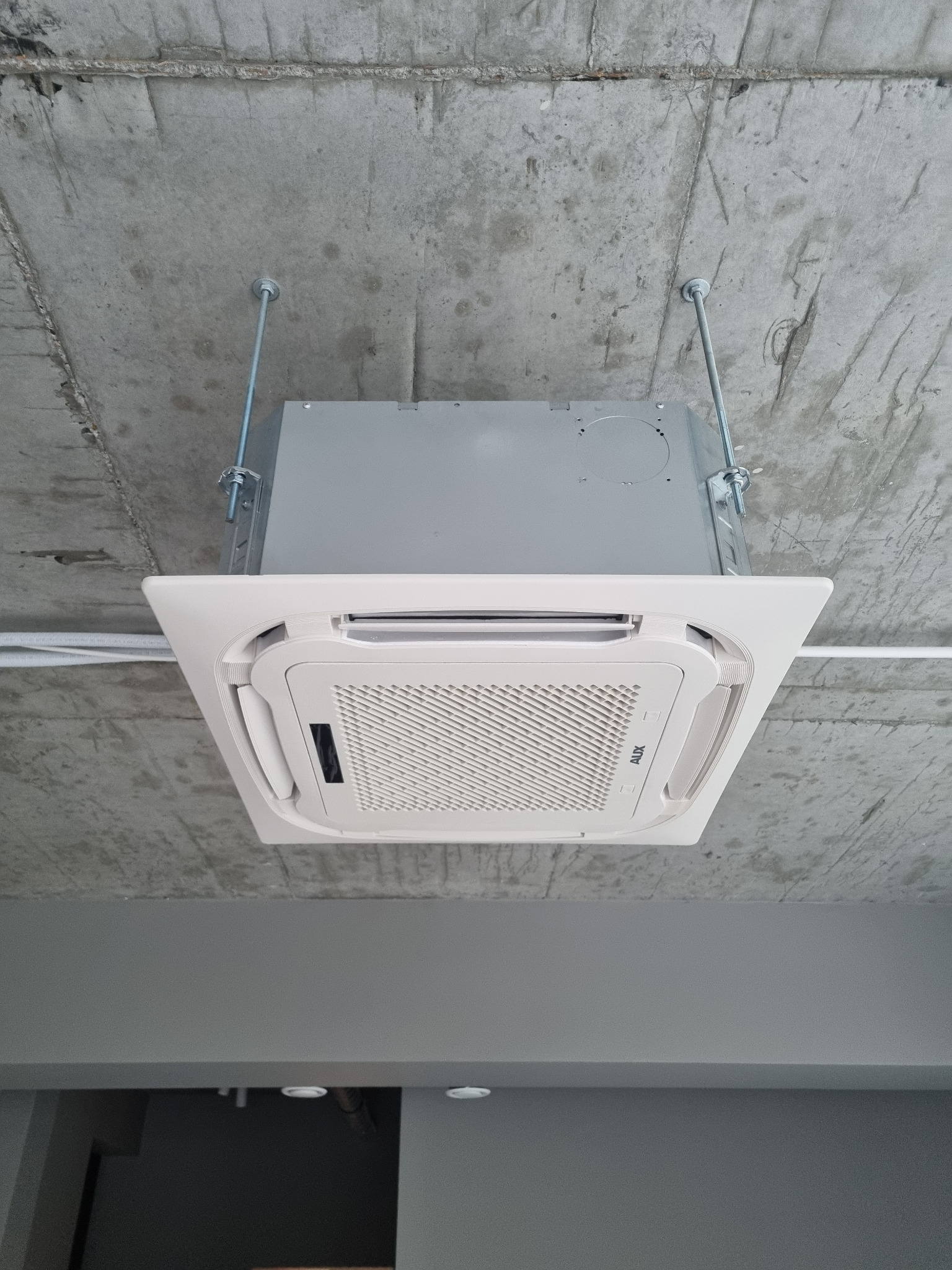 Nowoczesna, biała klimatyzacja kasetonowa AUX zamontowana pod surowym, betonowym sufitem. Widoczne metalowe pręty mocujące i biała instalacja elektryczna.