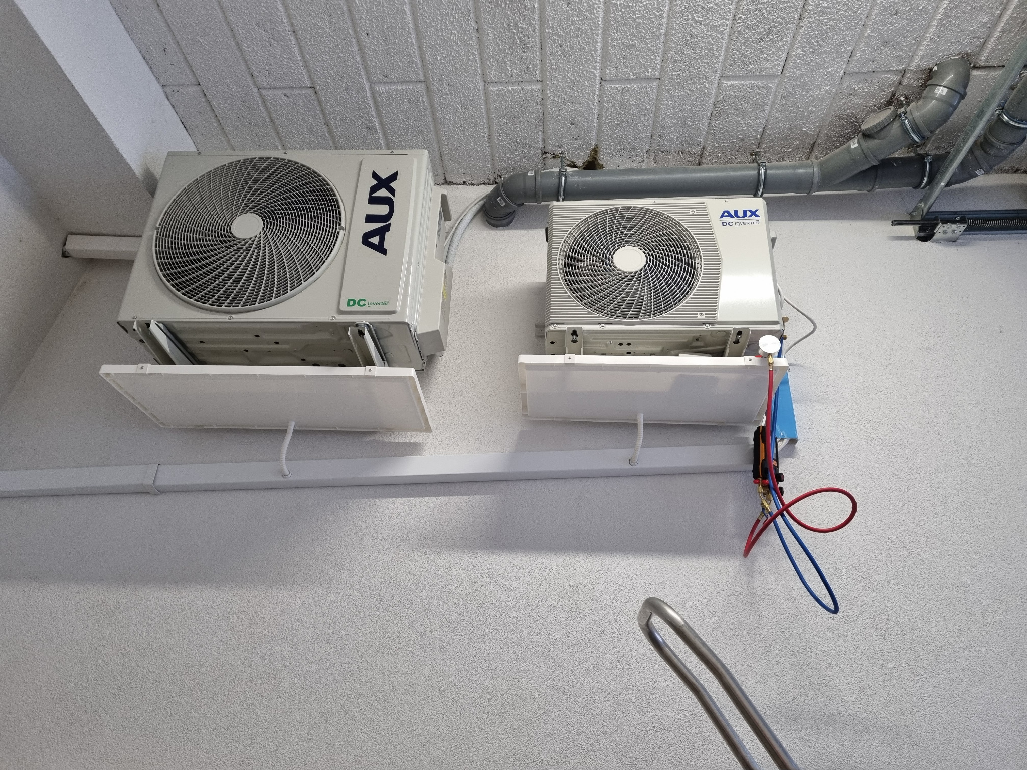 Dwa białe klimatyzatory AUX zamontowane na ścianie, z widocznymi przewodami instalacyjnymi i rurami wentylacyjnymi pod sufitem. Widok z dołu.