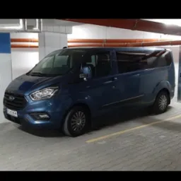 Niebieski Ford Transit Custom, zaparkowany w garażu podziemnym. Widoczne metalowe rury pod sufitem i szara kostka brukowa na podłodze. Tablica rejestracyjna widoczna.