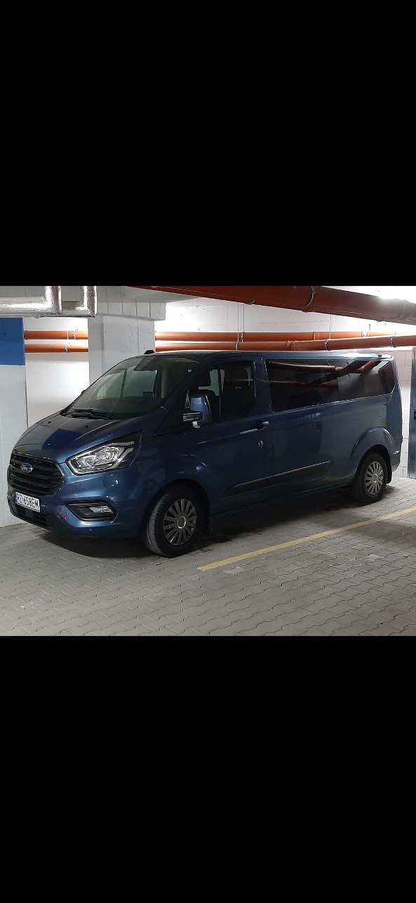 Niebieski Ford Transit Custom, zaparkowany w garażu podziemnym. Widoczne metalowe rury pod sufitem i szara kostka brukowa na podłodze. Tablica rejestracyjna widoczna.