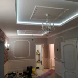 Wykończenie wnętrza z oświetleniem LED w suficie, dekoracyjnymi panelami ściennymi i ceglaną ścianą. W tle widoczne narzędzia i materiały budowlane.