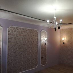 INSIDE_HOME SPÓŁKA Z OGRANICZONĄ ODPOWIEDZIALNOŚCIĄ - Eleganckie wnętrze po tapetowaniu: ściany w kwiatowe wzory, ozdobne listwy, kinkiety i żyrandol. Widoczny fragment osoby w lewym dolnym rogu.