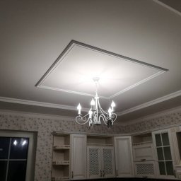 INSIDE_HOME SPÓŁKA Z OGRANICZONĄ ODPOWIEDZIALNOŚCIĄ - Wykończony sufit z dekoracyjną ramą i żyrandolem w kuchni z tapetą w kwiaty. Widok na zabudowę kuchenną w tle.