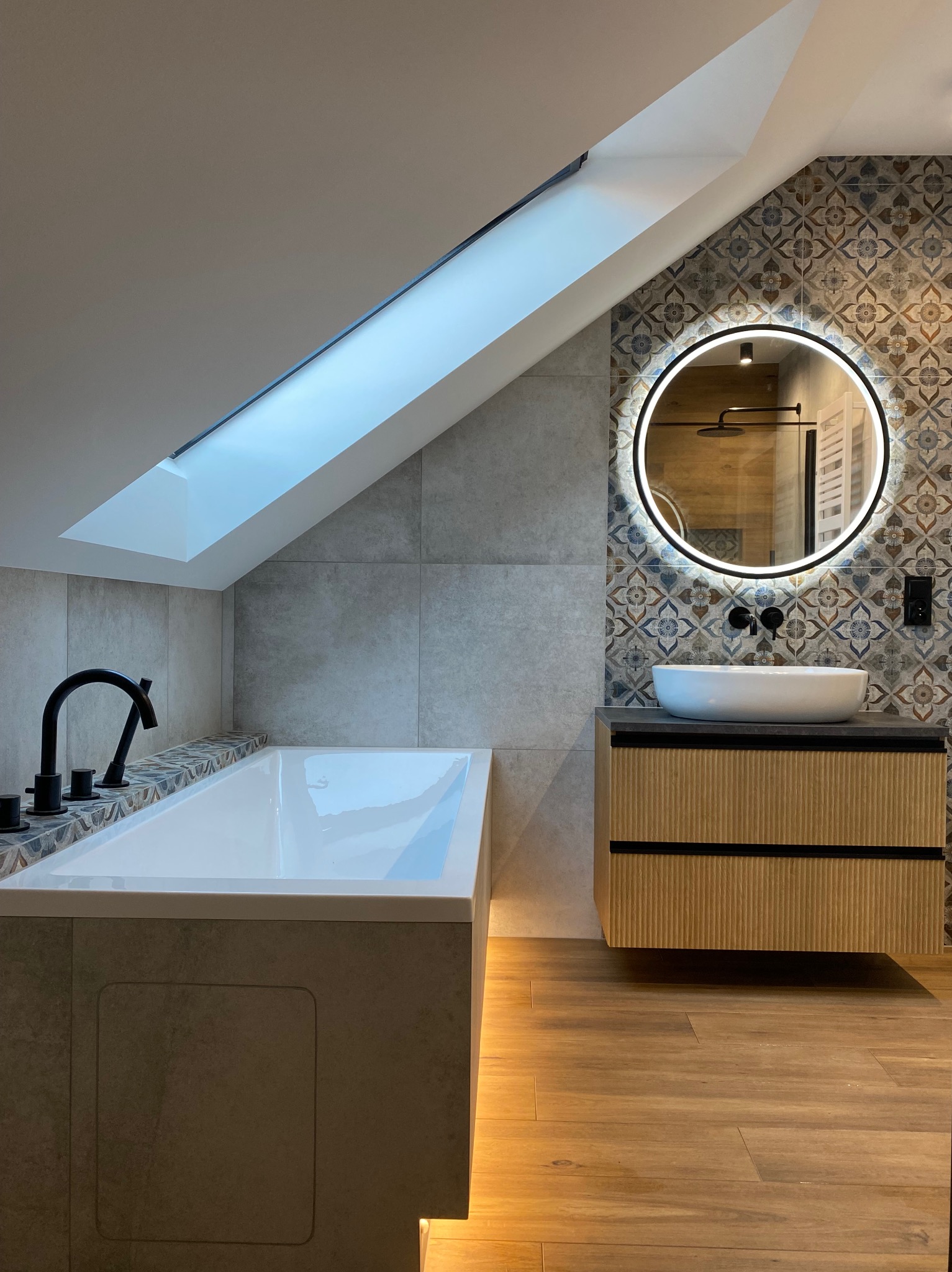 Nowoczesna łazienka na poddaszu z wanną obudowaną płytkami, okrągłym lustrem z podświetleniem i drewnianą szafką pod umywalką, wykończona w szarościach i brązach.