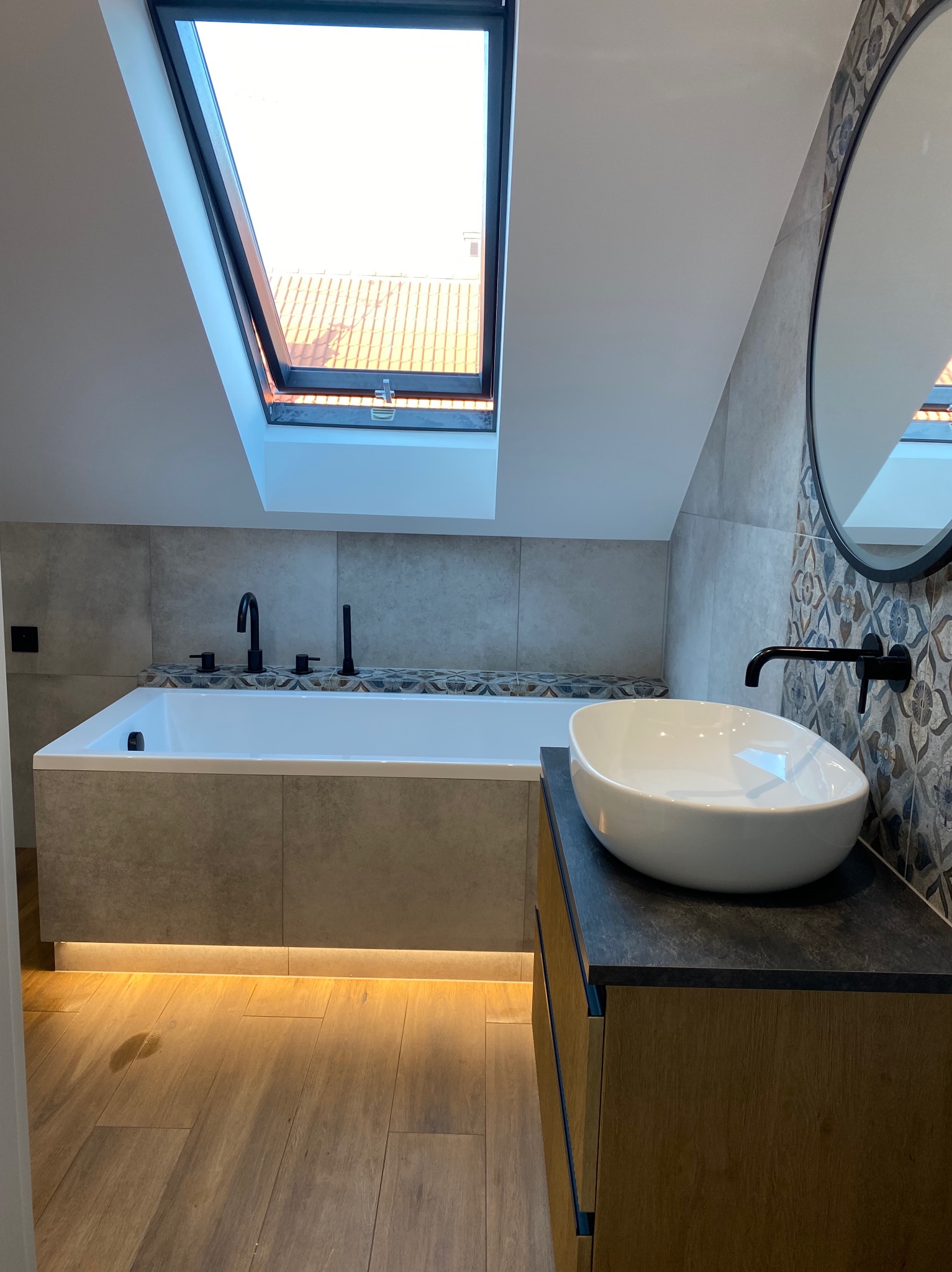 Stylowa łazienka na poddaszu z oknem dachowym, białą owalną umywalką na szafce z drewnopodobnym frontem i podświetleniem LED oraz wanną z czarną armaturą.