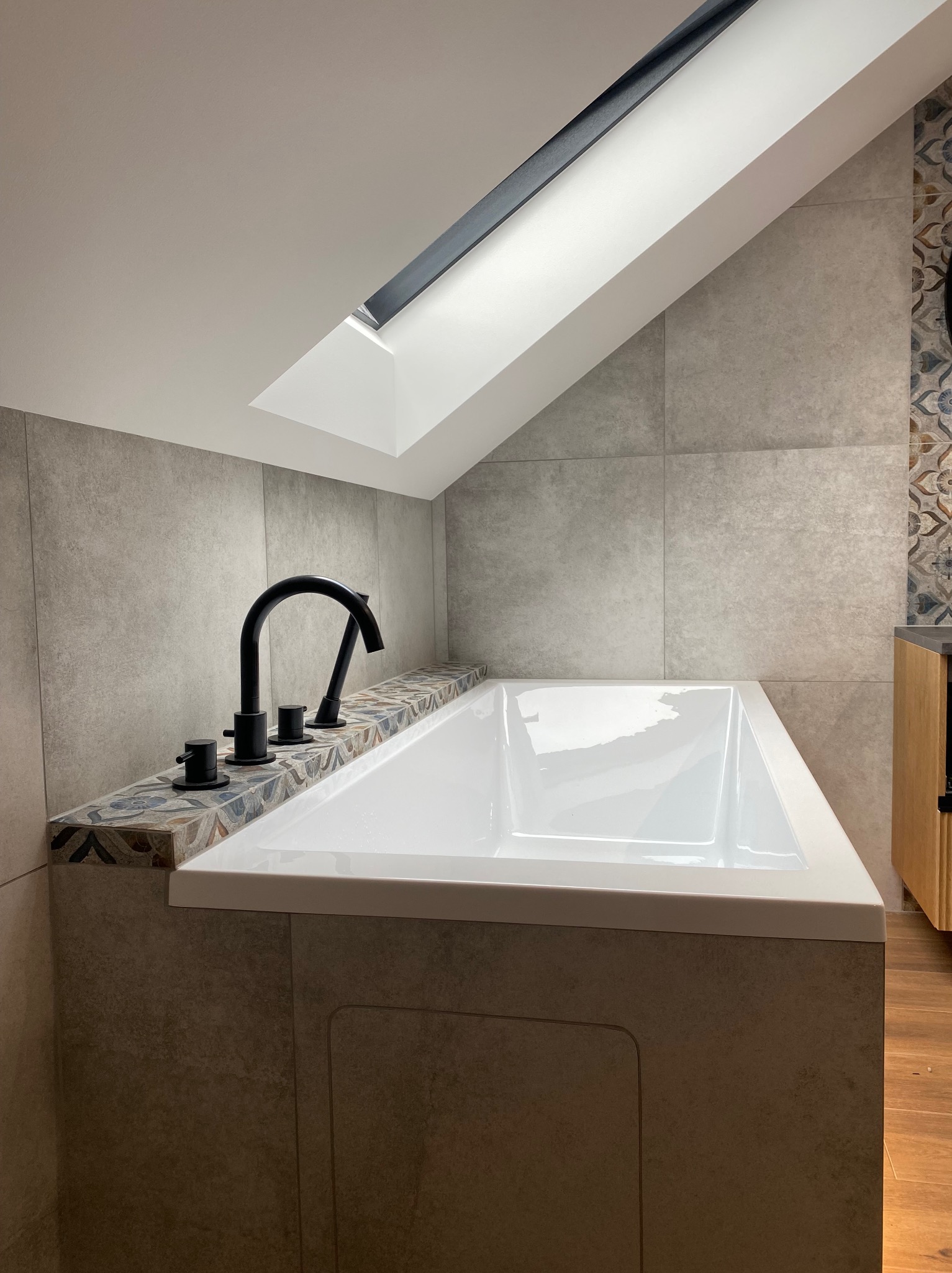 Nowoczesna wanna z czarną armaturą, obudowana płytkami imitującymi kamień, z dekoracyjnym pasem płytek. W tle szare płytki ścienne i okno dachowe.