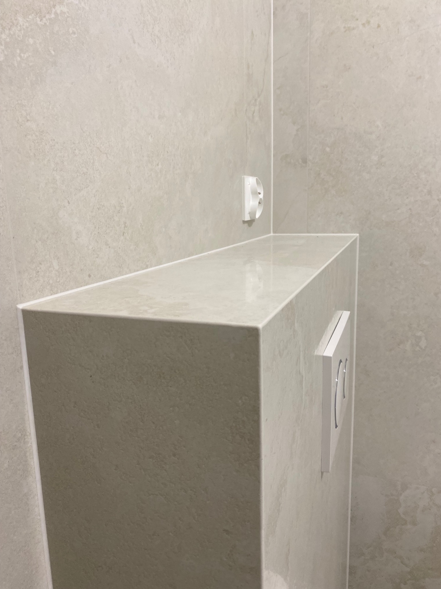 Obudowa stelaża WC wykończona płytkami w łazience, z widocznym przyciskiem spłukującym i gniazdkiem elektrycznym. Jasna kolorystyka, minimalistyczny design.