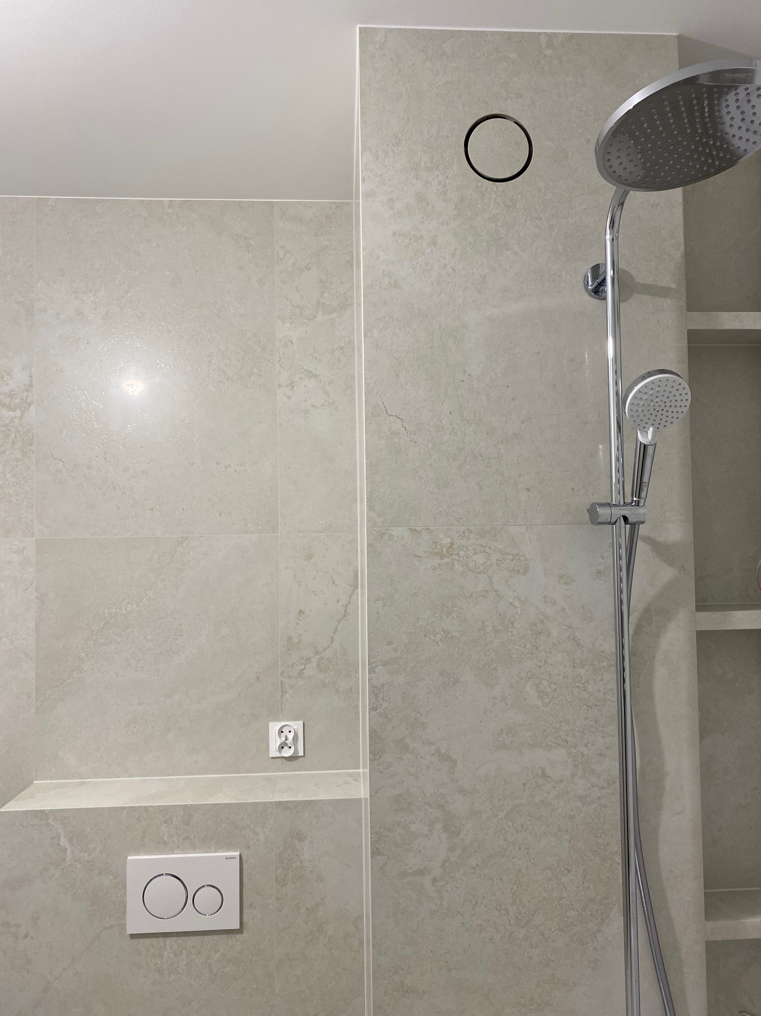 Wykończona kabina prysznicowa z beżowymi płytkami imitującymi kamień, chromowaną armaturą i wnęką z półkami. Widoczne gniazdko elektryczne i panel spłuczki.