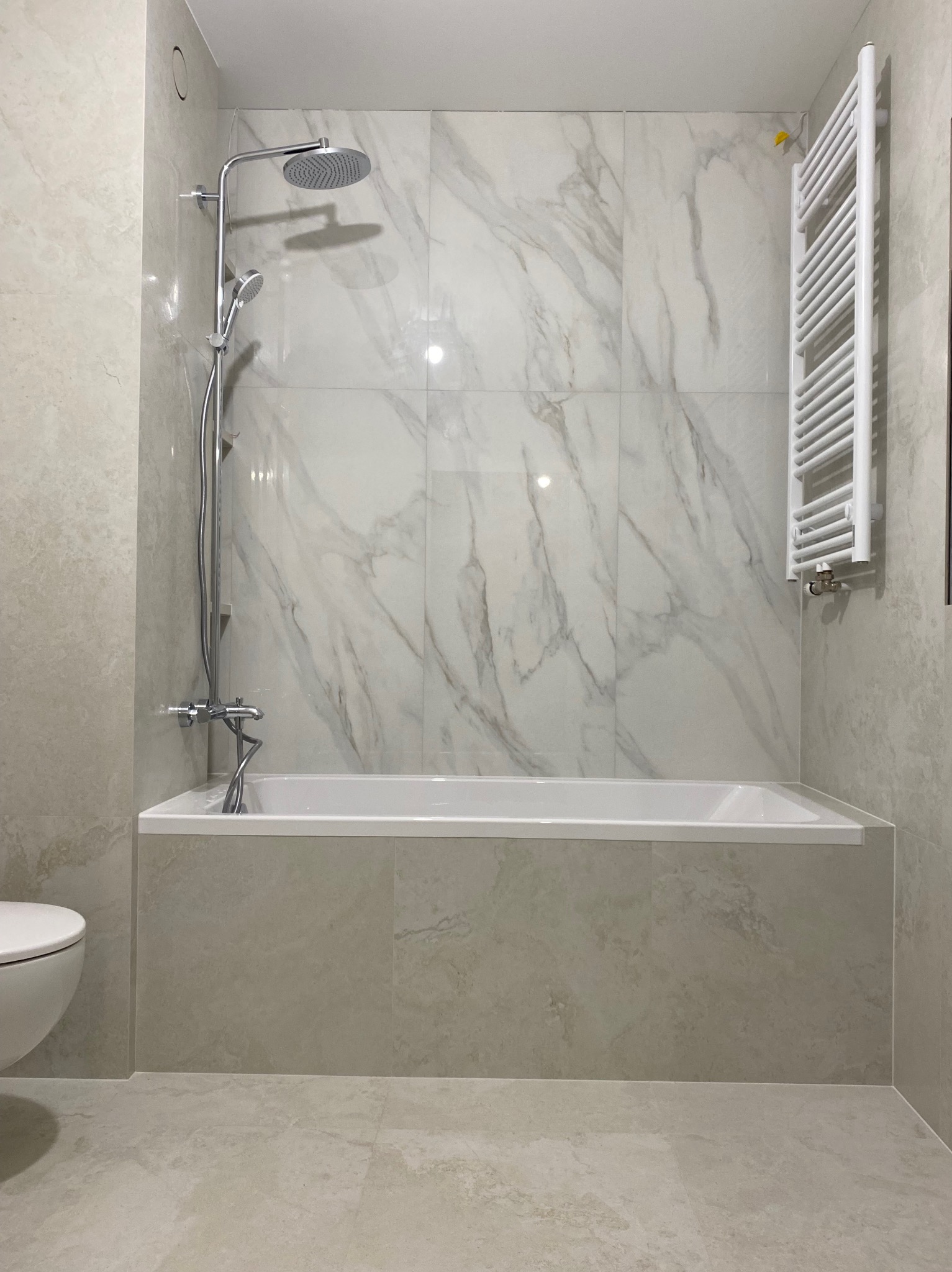 Nowoczesna łazienka z wanną obłożoną beżowymi płytkami imitującymi kamień, chromowaną armaturą i białym grzejnikiem drabinkowym.