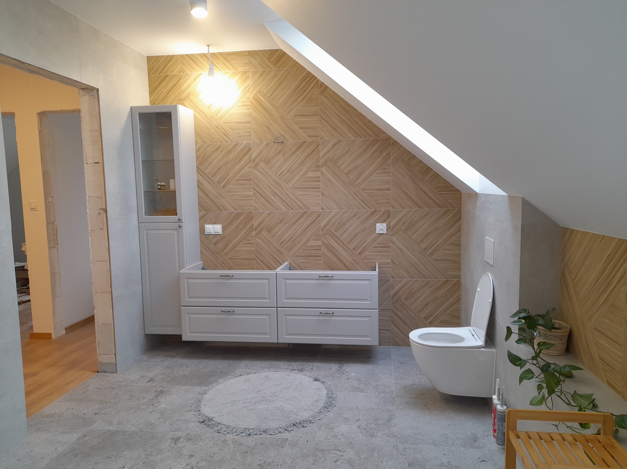 Jasna łazienka na poddaszu z drewnopodobnymi płytkami na ścianie, szarą podłogą i białymi meblami w trakcie montażu. Widoczny fragment otwartego przejścia.