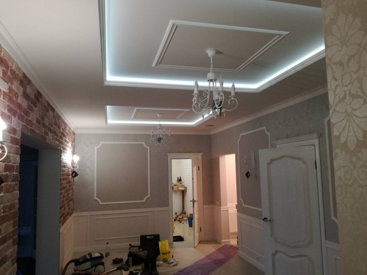 Wykończenie wnętrza z oświetleniem LED w suficie, dekoracyjnymi panelami ściennymi i ceglaną ścianą. W tle widoczne narzędzia i materiały budowlane.