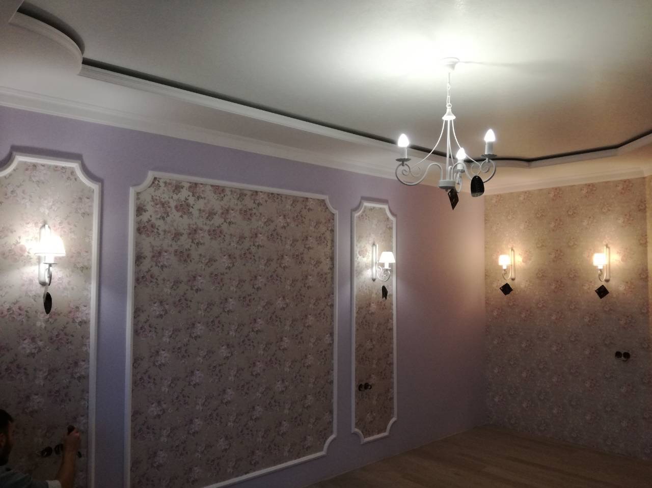 Eleganckie wnętrze po tapetowaniu: ściany w kwiatowe wzory, ozdobne listwy, kinkiety i żyrandol. Widoczny fragment osoby w lewym dolnym rogu.