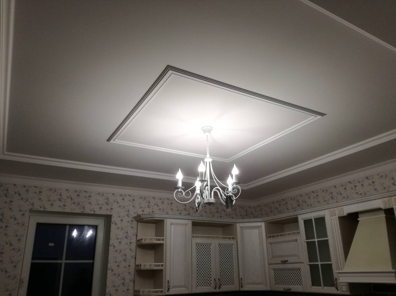 Wykończony sufit z dekoracyjną ramą i żyrandolem w kuchni z tapetą w kwiaty. Widok na zabudowę kuchenną w tle.