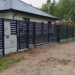STREFA OGRODZEŃ - Nowoczesne, poziome ogrodzenie metalowe w kolorze grafitowym, przylegające do budynku z numerem 37. Tło stanowią drzewa i częściowo widoczny pojazd.