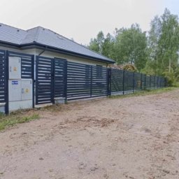 STREFA OGRODZEŃ - Nowoczesne, poziome ogrodzenie metalowe w kolorze antracytowym, otaczające posesję z domem. Widoczna brama wjazdowa i skrzynki elektryczne na słupku. W tle las.
