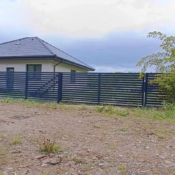 STREFA OGRODZEŃ - Nowoczesne, poziome ogrodzenie panelowe w kolorze antracytowym, otaczające posesję z domem jednorodzinnym. Widok z zewnątrz działki, w tle zieleń i pochmurne niebo.