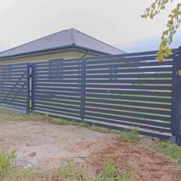 STREFA OGRODZEŃ - Nowoczesne, poziome ogrodzenie metalowe w kolorze grafitowym, z widocznymi przęsłami i słupkami, na tle domu jednorodzinnego i zieleni. Widok z poziomu gruntu.