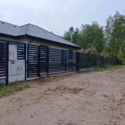 STREFA OGRODZEŃ - Nowoczesne, poziome ogrodzenie panelowe w kolorze antracytowym, z widocznymi skrzynkami elektrycznymi na tle domu jednorodzinnego i drzew. Widoczny samochód dostawczy w tle.