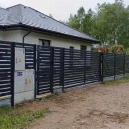 Nowoczesne, poziome ogrodzenie panelowe w kolorze antracytowym, z widoczną skrzynką elektryczną na słupku, na tle domu z szarą dachówką i zieleni.