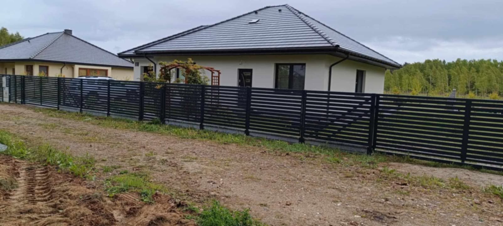 Nowoczesne, poziome ogrodzenie metalowe w kolorze czarnym przed domem z szarą dachówką. W tle drzewa i drugi budynek. Widoczne ślady opon na ziemi.