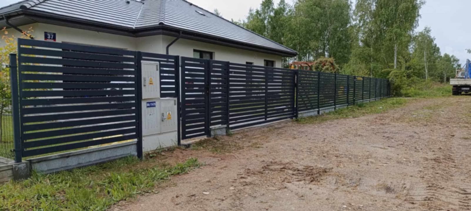 Nowoczesne, poziome ogrodzenie metalowe w kolorze grafitowym, przylegające do budynku z numerem 37. Tło stanowią drzewa i częściowo widoczny pojazd.