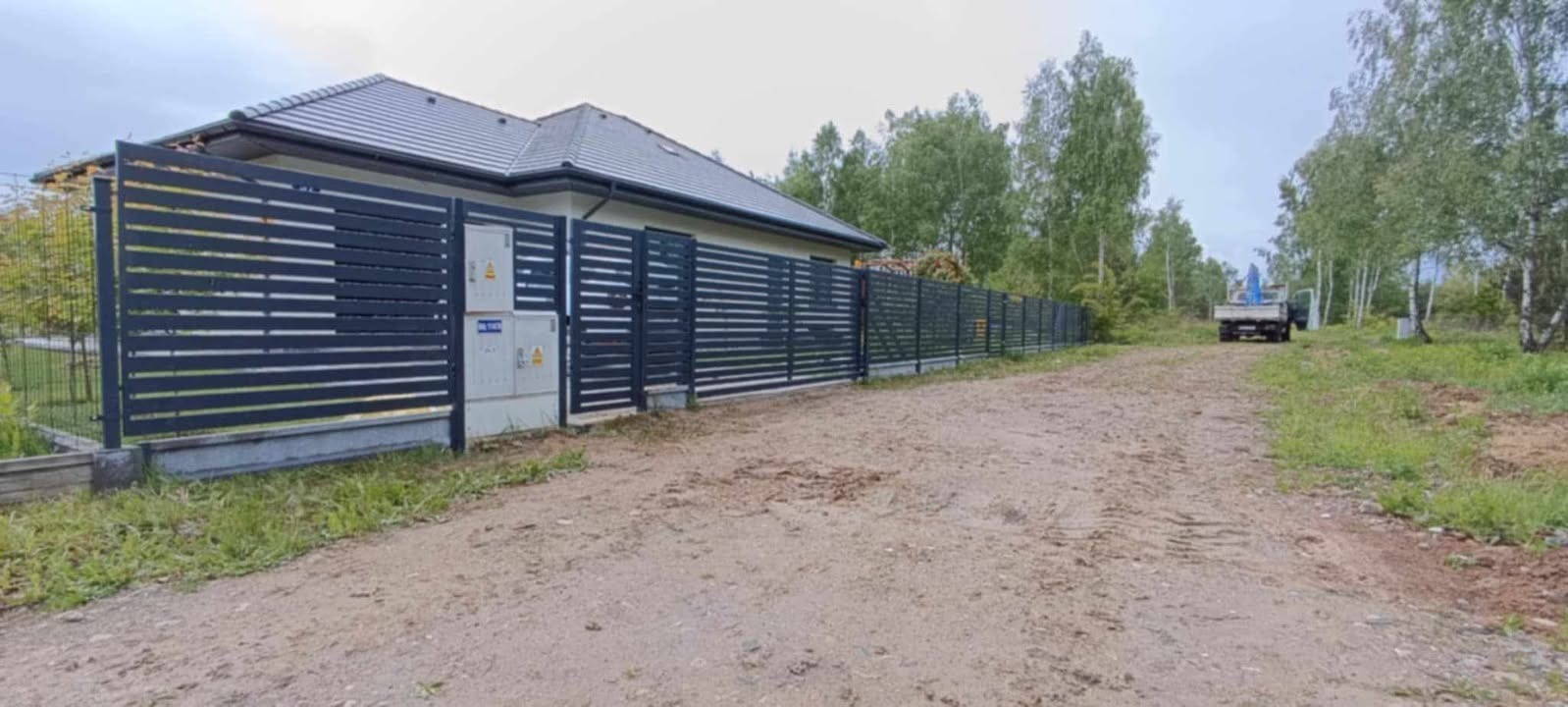 Nowoczesne, poziome ogrodzenie metalowe w kolorze antracytowym, otaczające posesję z domem. Widoczna brama wjazdowa i skrzynki elektryczne na słupku. W tle las.