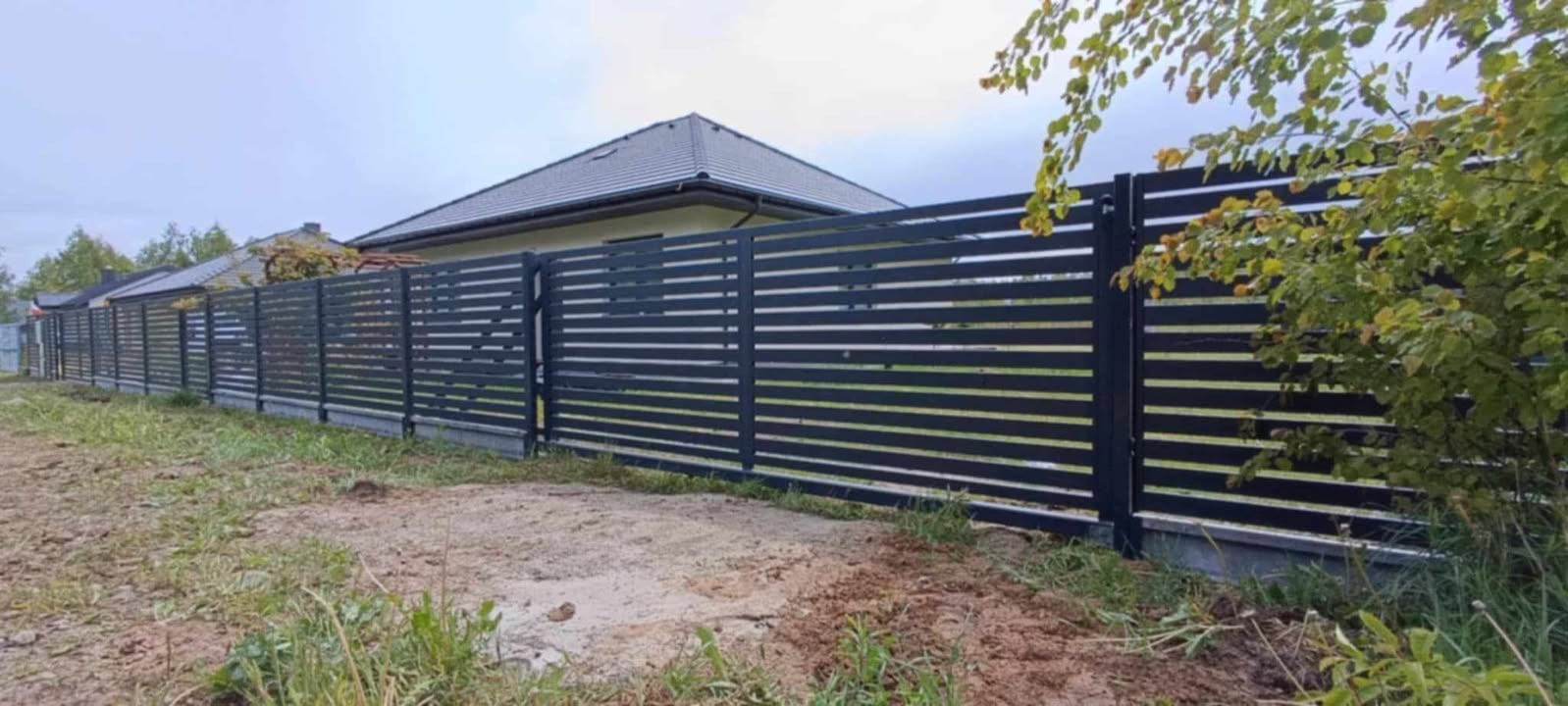 Nowoczesne, poziome ogrodzenie metalowe w kolorze antracytowym, z bramą. Tło to dom z szarą dachówką i zieleń. Minimalistyczny design, świeżo posadzona trawa.