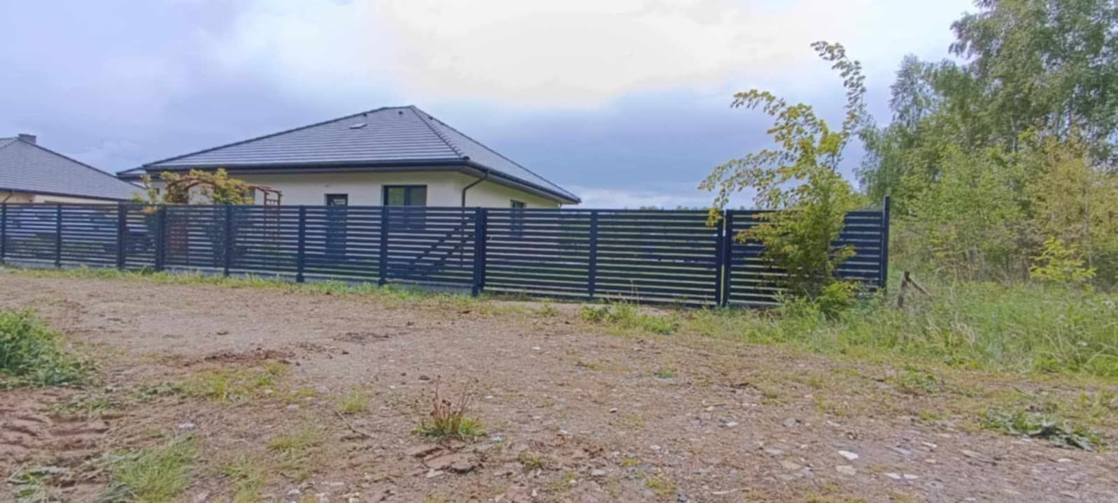 Nowoczesne, poziome ogrodzenie panelowe w kolorze antracytowym, otaczające posesję z domem jednorodzinnym. Widok z zewnątrz działki, w tle zieleń i pochmurne niebo.