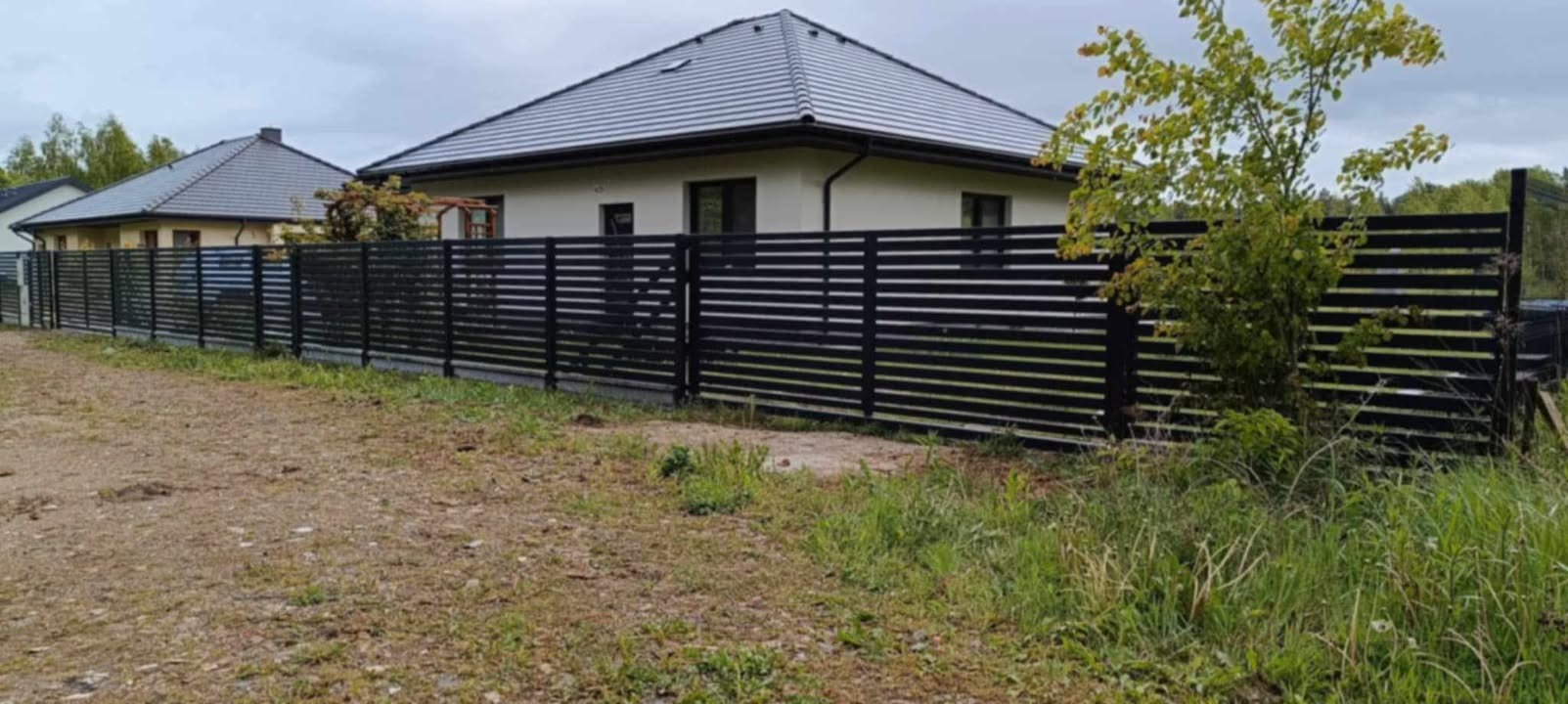 Nowoczesne, poziome ogrodzenie metalowe w kolorze czarnym, z widocznym domem w tle. Minimalistyczny design i staranne wykonanie.