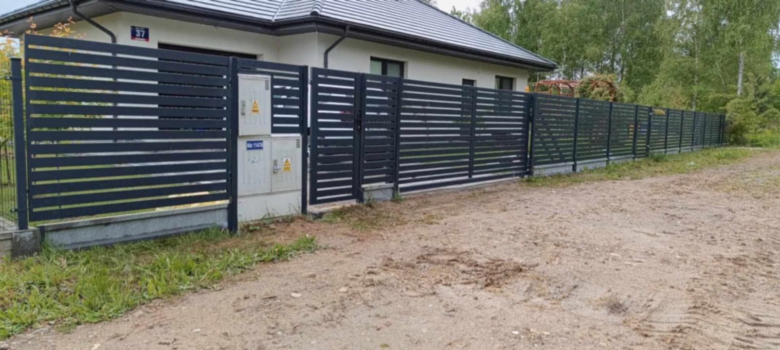 Nowoczesne, poziome ogrodzenie panelowe w kolorze antracytowym, z numerem domu 37, na tle domu z jasną elewacją i zieleni. Widoczna skrzynka elektryczna.