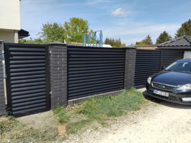 Nowoczesne, poziome, czarne ogrodzenie z ceglanymi słupkami, brama wjazdowa i zaparkowany samochód osobowy. Ujęcie z zewnątrz posesji.