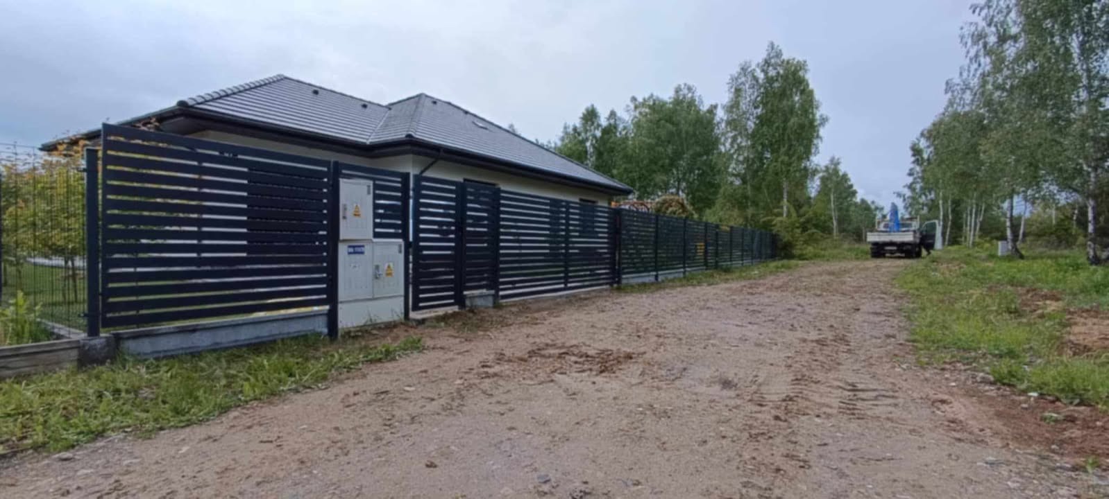 Nowoczesne, poziome ogrodzenie panelowe w kolorze antracytowym, z widocznymi skrzynkami elektrycznymi na tle domu jednorodzinnego i drzew. Widoczny samochód dostawczy w tle.