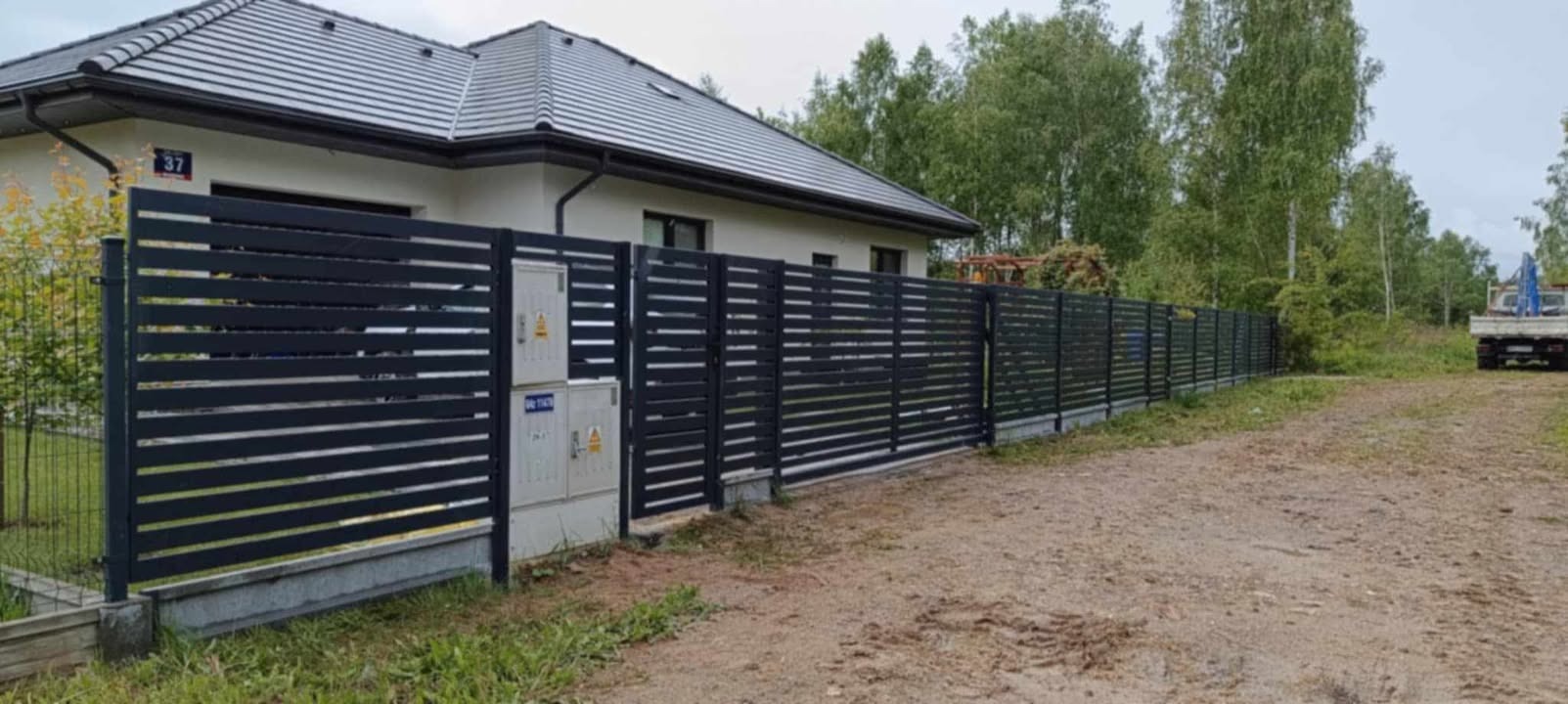 Nowoczesne, poziome ogrodzenie panelowe w kolorze antracytowym, z widoczną skrzynką elektryczną na słupku, na tle domu z szarą dachówką i zieleni.