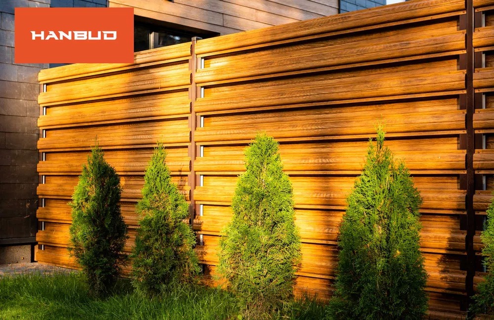 Nowoczesne ogrodzenie z drewnopodobnych paneli w ciepłym odcieniu brązu, z zielonymi tujami. Widoczne elementy metalowej konstrukcji. Logo firmy w lewym górnym rogu.