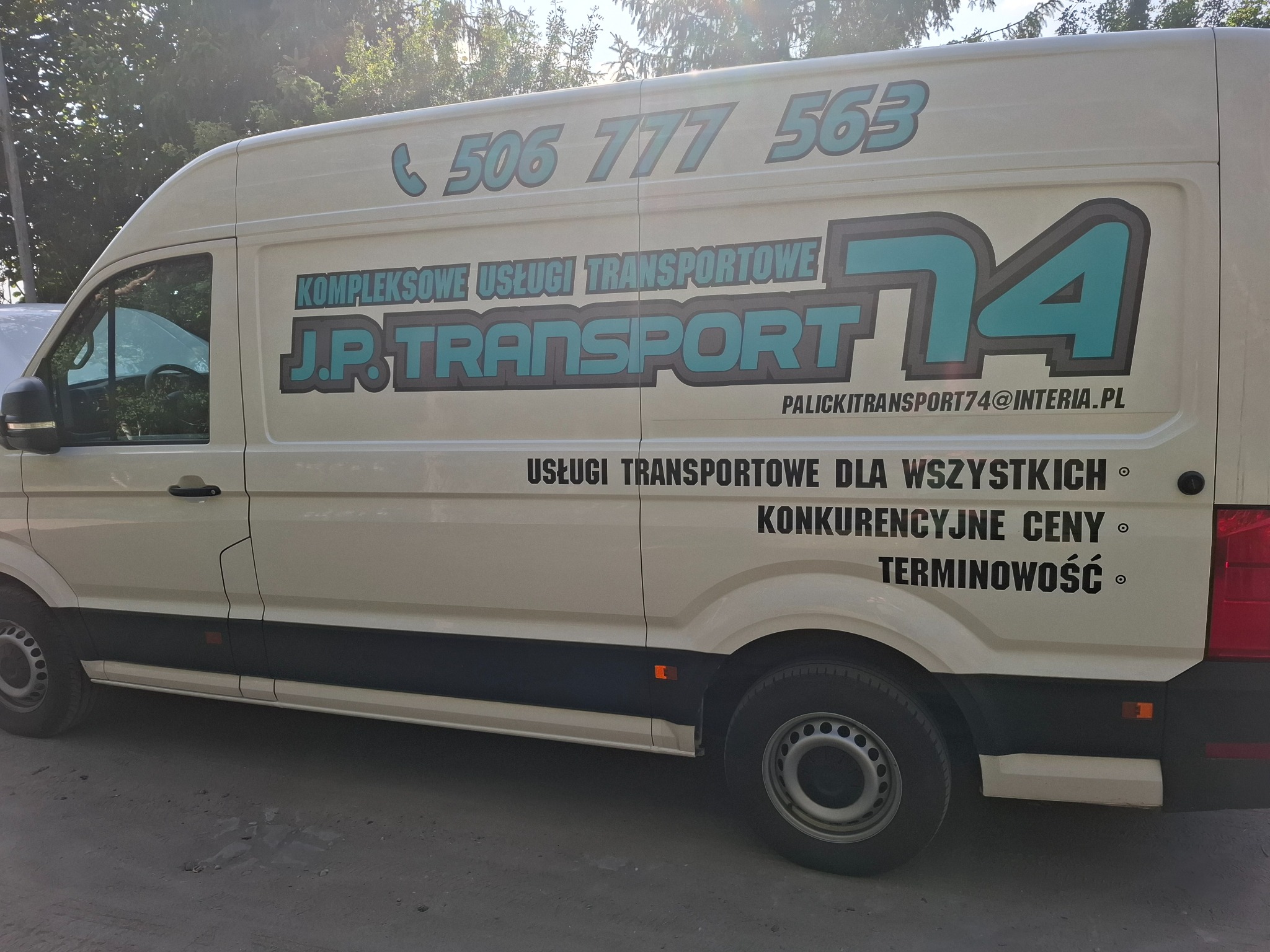 Biały bus z niebieskim logo J.P. Transport 74, numerem telefonu i hasłami reklamowymi na boku, zaparkowany na szarej nawierzchni, widok z boku w słoneczny dzień.