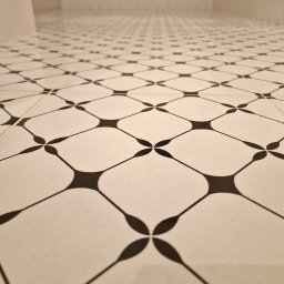 USŁUGI REMONTOWO-WYKOŃCZENIOWE ABIG-BUD GRZESIAK ADRIAN - Białe płytki podłogowe z czarnym wzorem w kształcie kwiatów, ułożone w łazience. Perspektywa z dołu, widoczny fragment ściany w tle.