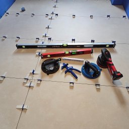 USŁUGI REMONTOWO-WYKOŃCZENIOWE ABIG-BUD GRZESIAK ADRIAN - Ułożone płytki podłogowe z systemem poziomowania, poziomice, przyssawki do płytek i narzędzia glazurnicze w trakcie pracy. Widok z góry na podłogę w niebieskim pomieszczeniu.