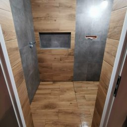 USŁUGI REMONTOWO-WYKOŃCZENIOWE ABIG-BUD GRZESIAK ADRIAN - Wykończona łazienka z drewnopodobnymi i szarymi płytkami, wnęką w ścianie i widoczną instalacją elektryczną. Widok zza otwartych drzwi.