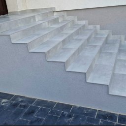 USŁUGI REMONTOWO-WYKOŃCZENIOWE ABIG-BUD GRZESIAK ADRIAN - Zewnętrzne schody wyłożone szarymi płytkami, z subtelnymi rowkami antypoślizgowymi. Ciemna kostka brukowa u podstawy i obok schodów. Elewacja budynku w odcieniach szarości.